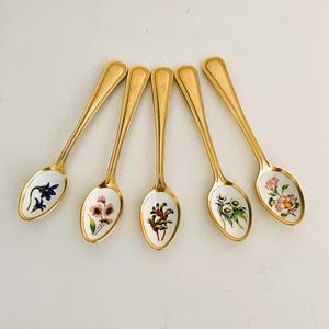 May include: Five gold-coloured spoons with floral designs in the bowls. Each spoon has a different flower painted in the bowl, with gold detailing on the handle and rim. The spoons are arranged in a fan shape.