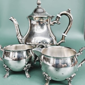 May include: A silver tea set with intricate designs. The set comprises a teapot, a creamer, and a sugar bowl. The teapot features a curved spout and a decorative handle. The creamer and sugar bowl have small handles and rest on decorative feet. The set is displayed on a green surface.