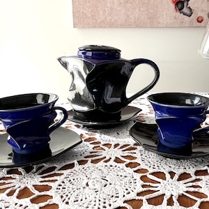 May include: A contemporary tea set comprising a black and blue teapot, cups, and saucers. The cups and teapot showcase a distinctive, twisted design. The set is presented on a white lace tablecloth, offering a contrast of textures and colours.