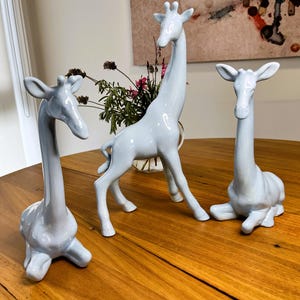 May include: Three ceramic giraffe figurines in a light blue colour. The giraffes are in different poses, with one standing tall, one sitting down, and one standing with its head tilted to the side. The figurines are on a wooden table.