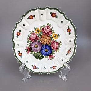 May include: A round ceramic plate with a scalloped edge and a dark green rim. The centre features a colourful hand-painted floral bouquet with pink, blue, purple, and orange flowers. Small floral motifs decorate the fluted border of the plate.