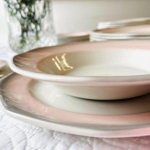 May include: A set of white ceramic dinnerware with a pink and grey rim. The plates are stacked on top of each other, with a bowl on top.
