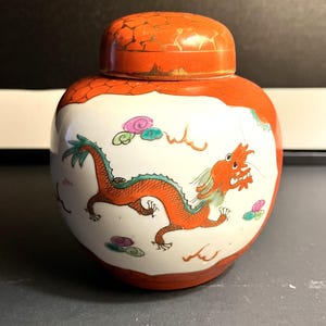 May include: A round ceramic jar with a lid, featuring a dragon design. The jar is white with an orange dragon and a matching orange lid. The dragon is red and green, with pink and blue accents. The lid has a cracked orange finish.