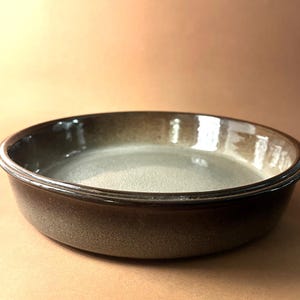 May include: A round ceramic baking dish with a dark brown exterior and a lighter, speckled interior. The dish has a slightly raised rim and a smooth, glossy finish. The overall colour palette is earthy and neutral.