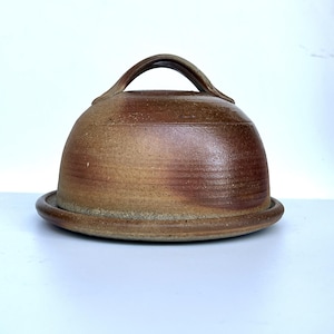 May include: A handmade, brown ceramic cheese dome with a matching base. The dome has a curved handle on top. The pottery has a rustic, earthy appearance, with variations in colour and texture. The base is a simple, round plate.