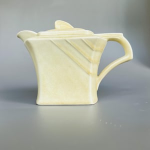May include: A pale yellow ceramic creamer featuring a square-shaped body with angled geometric lines on the upper sides, a small spout, a curved handle, and a matching lid with a triangular finial, showcasing a smooth, glossy finish.
