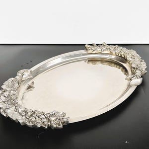 May include: An oval, silver-plated serving tray with floral garland handles. The tray has a polished surface and is decorated with detailed, three-dimensional flowers and leaves. The handles are formed by the floral design, adding a decorative touch to the serving piece.