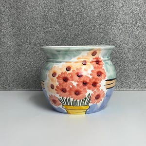 May include: A ceramic planter featuring a hand-painted design of orange flowers in a yellow pot. The background of the planter is decorated with soft blue and green watercolour-style washes, and the rim is finished with a light green painted band.