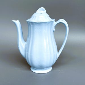 May include: A light blue ceramic teapot featuring a vertical ribbed pattern, a curved handle, a pointed spout, and a matching lid with a small finial. The surface of the teapot displays a visible network of fine cracks throughout the glaze.