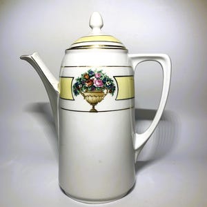 May include: White ceramic coffee pot with a lid and handle. The pot has a yellow band with a floral design and gold trim. The spout is on the left, and the handle is on the right.