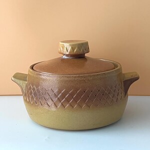 May include: A round, lidded ceramic casserole dish in shades of mustard yellow and brown. The body features a textured, diamond-shaped lattice pattern around the centre, two side handles, and a matching lid with a small, knob-like handle.