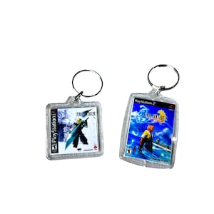 Może przedstawiać: Dwa przezroczyste breloki z plastikowymi obrazkami gry wideo Final Fantasy VII i Final Fantasy IX. Breloki są kwadratowe i mają logo PlayStation.