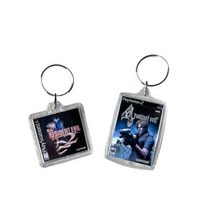 May include: Two keychains featuring video game covers. The first keychain is silver and features the Resident Evil 2 cover art with the PlayStation logo. The second keychain is silver and features the Resident Evil 4 cover art with the PlayStation 2 logo.