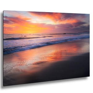 May include: A vibrant beach scene at sunset. The sky is filled with orange, pink, and purple hues, reflecting on the wet sand and ocean waves. The composition captures the serene beauty of the coastal landscape.
