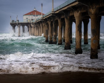May include: A concrete pier extends over a choppy ocean with large waves crashing against the pillars. The pier has a white building with a red roof at the end.
