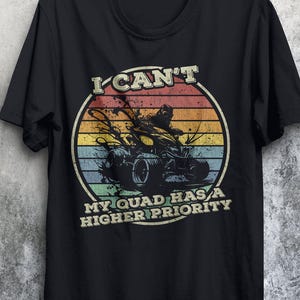 May include: Black t-shirt with a vintage-style graphic of a person riding a quad. The graphic has a sunset background and the text "I can't my quad has a higher priority".