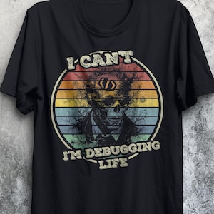 Könnte beinhalten: Schwarzes T-Shirt mit einem Vintage-Grafik eines Schädels mit Code in seinen Augen. Die Grafik ist von einem regenbogenfarbenen Sonnenuntergang umgeben und dem Text "Ich kann nicht, ich debugge das Leben".