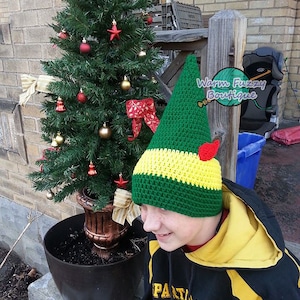 May include: A green and yellow crocheted elf hat with a red pom-pom on top. The hat is worn by a person with a yellow and black hoodie. The text "Warm Fuzzy Boutique" is visible in the image.