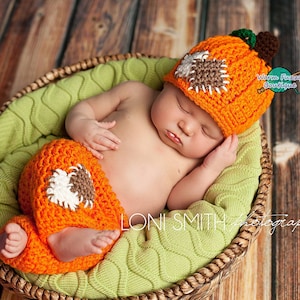 May include: A newborn baby wearing a crocheted pumpkin hat and matching pants. The baby is sleeping in a wicker basket lined with green fabric.