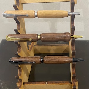 May include: Three wooden pens with different coloured wood and metal accents are displayed on a wooden stand. The pens are arranged vertically on the stand.