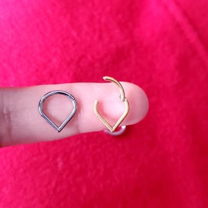 May include: Two teardrop-shaped nose rings, one silver and one gold, are shown on a finger. The rings are open and ready to be worn.