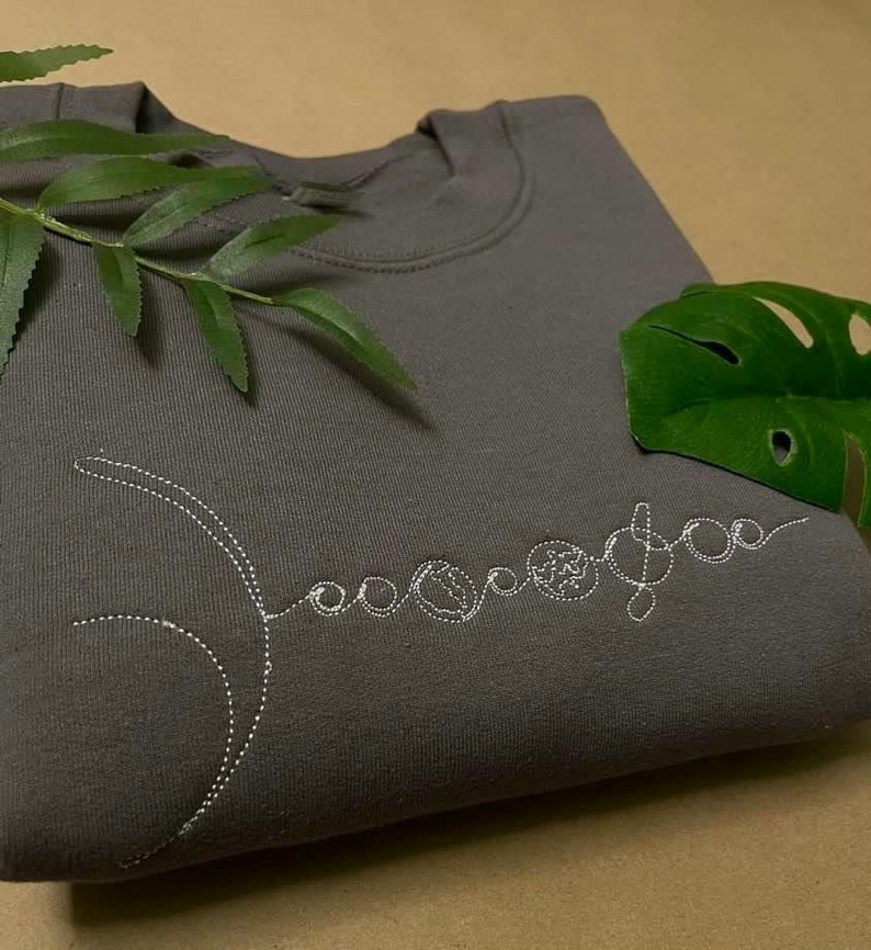 Puede incluir: Una sudadera gris con un dise&ntilde;o bordado blanco de una luna creciente y planetas. El dise&ntilde;o est&aacute; cosido en un estilo de l&iacute;nea punteada.
