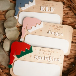 Peut inclure: Trois pancartes en bois avec des espaces vierges pour écrire des messages. Chaque pancarte présente un chapeau d'elfe différent en rouge, rose et bleu avec une couronne blanche. Les pancartes portent le texte "A message from your Elf", "A message from Jingle", et "A message from Sprinkles".
