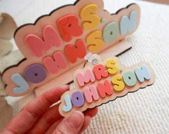 May include: A wooden sign with the text "Mrs. Johnson" in colorful, puffy letters. The sign is shaped like a cloud and has a small loop for hanging.