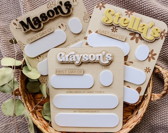May include: Three wooden signs with blank spaces for writing. The signs are decorated with a floral pattern and have the names "Mason's", "Stella's", and "Grayson's" in a retro font. The signs are for a first day of school or last day of school celebration.