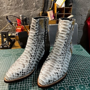 Puede incluir: Botines con estampado de piel de serpiente en tonos grises y blancos. Los botines tienen punta afilada, cremallera lateral y suela de cuero marrón. Se muestran sobre una esterilla de corte verde. Miden unos 20 cm de alto.