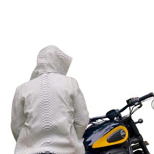 May include: A white, textured hooded jacket with a snakeskin pattern. The jacket is worn while seated on a black and yellow motorcycle. The motorcycle has a black seat and handlebars.