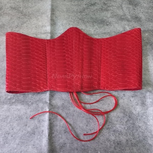 Könnte beinhalten: Ein leuchtend rotes Korsett mit Schlangenleder-Textur. Das Korsett hat ein strukturiertes Design mit vertikalen Bahnen und einem Schnürverschluss. Der Text "NDASPYTHON" ist auf der Vorderseite sichtbar.