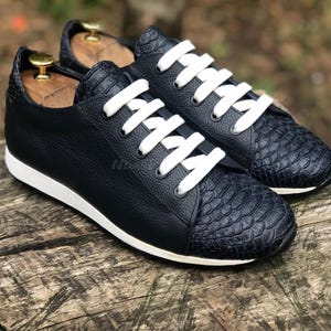 Pode incluir: Um par de tênis de couro azul marinho com cadarços brancos. A ponta do sapato tem um padrão de pele de cobra texturizado.