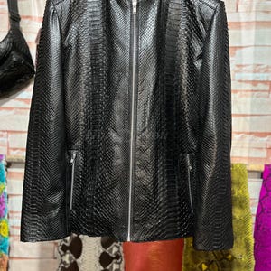 Puede incluir: Chaqueta negra de piel sintética con estampado de serpiente, con cuello alzado y cremallera completa. La chaqueta tiene dos bolsillos con cremallera y un patrón texturizado. Cuelga de una percha de madera.