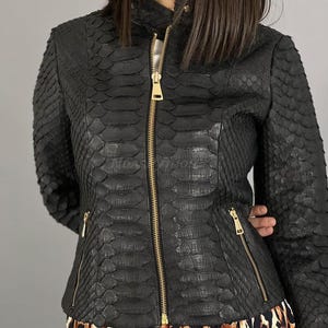 Pode incluir: Uma jaqueta de couro preta com padrão de pele de cobra e um zíper dourado. A jaqueta tem dois bolsos na frente e um bolso com zíper na manga esquerda.