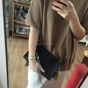 May include: Black clutch bag with a textured design, silver chain, and tassel. The bag is held by a person wearing a silver watch. A wooden shelf with handbags is visible in the background.