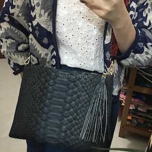 Puede incluir: Bolso de mano negro con textura de piel de serpiente y borla a juego. El bolso tiene una cremallera dorada y una correa estampada en rojo y marrón. El texto "NdasPython" es visible en el bolso.