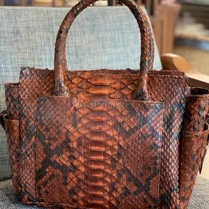 May include: A brown snakeskin handbag with a top handle. The bag has a textured snakeskin pattern with black and brown accents.
