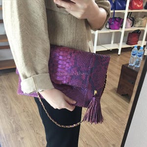 Puede incluir: Un bolso de mano morado con un estampado de piel de serpiente. El bolso tiene una correa de cadena dorada y una borla a juego. El texto "NdusPython" es visible en la parte delantera.