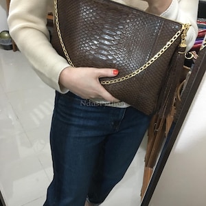 Może przedstawiać: Brązowa kopertówka ze wzorem skóry węża, ze złotym łańcuszkiem i zamkiem błyskawicznym. Torba ma frędzel i jest trzymana przez osobę ubraną w biały sweter i niebieskie dżinsy.