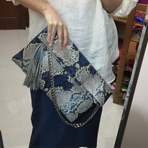 Könnte beinhalten: Eine rechteckige Clutch mit Schlangenmuster in Blau-, Grau- und Weißtönen. Sie hat einen silbernen Kettenriemen und eine passende Quaste. Die Clutch wird von einer Person gehalten.