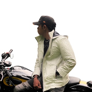 Op de afbeelding: Een crèmekleurige, getextureerde leren jas met ritssluiting en capuchon. De jas wordt over een zwart shirt gedragen. De persoon zit op een zwarte motorfiets met gele accenten. Een zwarte baseballpet wordt gedragen.