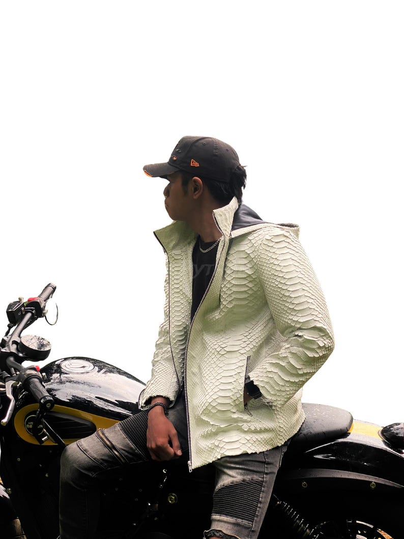 May include: A cream-coloured, textured leather jacket with a zip closure and a hood. The jacket is worn over a black shirt. The person is sitting on a black motorcycle with yellow accents. A black baseball cap is worn.