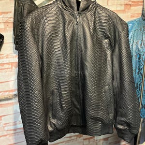 Puede incluir: Chaqueta negra con capucha y estampado de piel de serpiente. La chaqueta tiene cierre de cremallera, capucha y bolsillo delantero. Cuelga de una percha de madera.