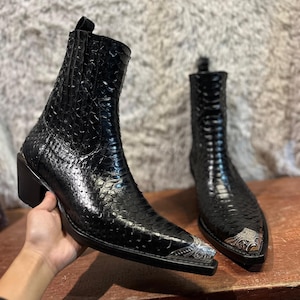 Puede incluir: Un par de botas de vaquero de cuero negro con textura de piel de serpiente. Las botas tienen puntas puntiagudas con detalles metálicos plateados y tacones cuadrados pequeños. Una bota está sujeta por una mano, la otra está sobre una superficie de madera.