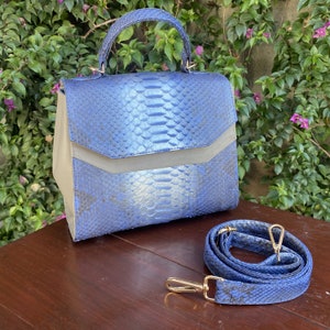 May include: A blue handbag with a snakeskin pattern, a top handle, and a detachable shoulder strap. The bag has a flap closure and gold-colored hardware. The bag is sitting on a wooden surface.
