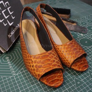 Puede incluir: Un par de sandalias de tacón abierto de cuero marrón con estampado de piel de serpiente. Las sandalias tienen una tira elástica negra en la parte superior del pie.