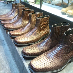 Peut inclure: Une rangée de bottes en cuir marron avec un motif en peau de serpent. Les bottes ont une pointe pointue et une fermeture éclair sur le côté.