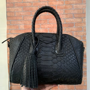 May include: A black leather handbag with a snakeskin pattern and a tassel. The bag has two top handles and a shoulder strap.