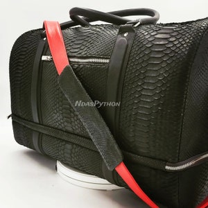 Può includere: Borsa da viaggio in pelle nera con motivo a pelle di serpente e tracolla rossa. La borsa ha una chiusura a zip e due manici superiori.